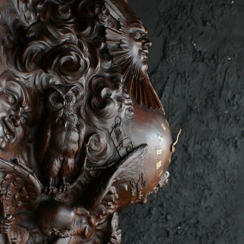 fig 3. A rectangular dark wood carved panel featuring a central heart-shaped clock face surrounded by allegorical figures, a sun motif, and clouds within a moulded wooden frame.