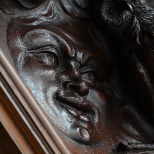 fig 4. A rectangular dark wood carved panel featuring a central heart-shaped clock face surrounded by allegorical figures, a sun motif, and clouds within a moulded wooden frame.