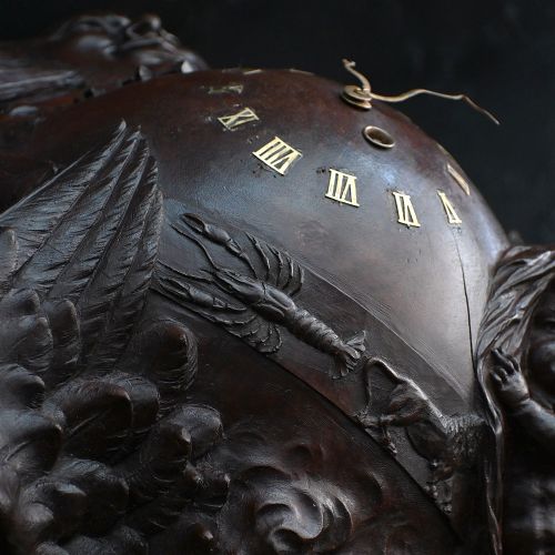 fig 7. A rectangular dark wood carved panel featuring a central heart-shaped clock face surrounded by allegorical figures, a sun motif, and clouds within a moulded wooden frame.