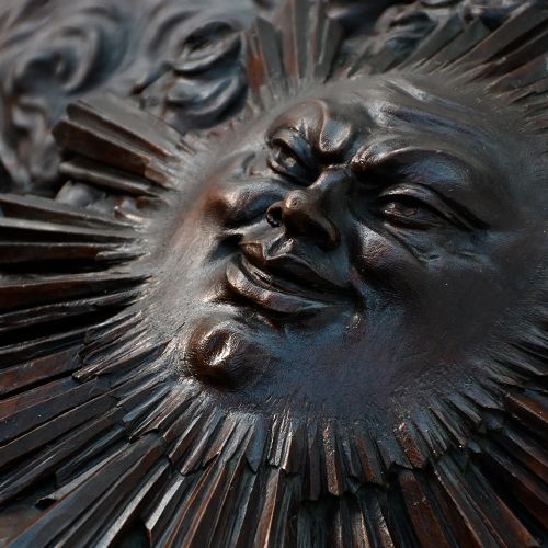 fig 11. A rectangular dark wood carved panel featuring a central heart-shaped clock face surrounded by allegorical figures, a sun motif, and clouds within a moulded wooden frame.
