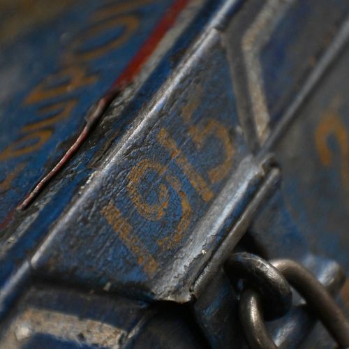 fig 2. An antique painted tin donation box featuring a Gallic cockerel standing atop a blue metal box inscribed with dates and fitted with a lock.