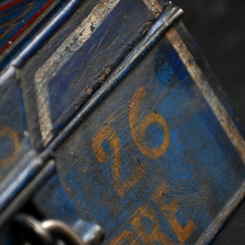 fig 3. An antique painted tin donation box featuring a Gallic cockerel standing atop a blue metal box inscribed with dates and fitted with a lock.