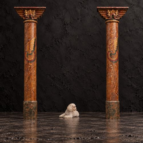 fig 2. A matched pair of early 20th-century tall wooden fairground carousel columns featuring flared carved capitals, faux-marble painted shafts with winged motifs, and dark green square plinths.