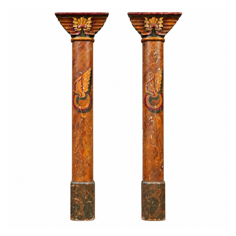 A matched pair of early 20th-century tall wooden fairground carousel columns featuring flared carved capitals, faux-marble painted shafts with winged motifs, and dark green square plinths.
