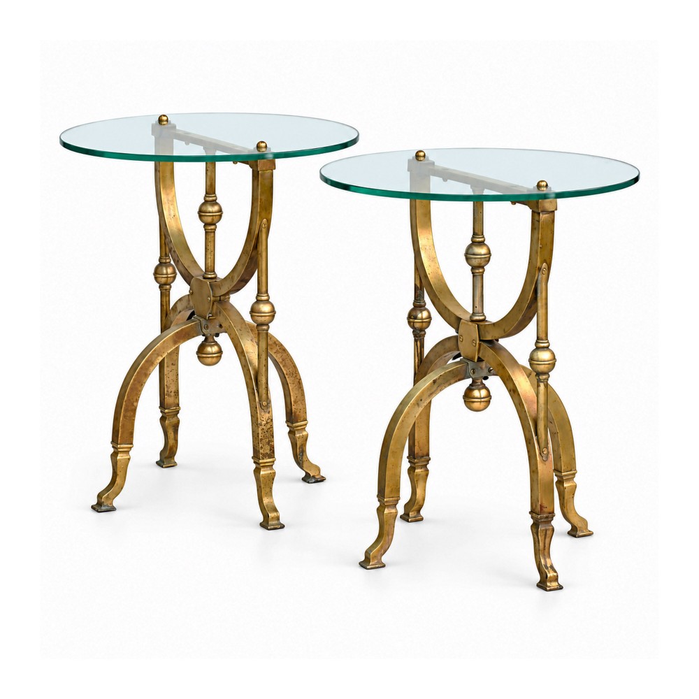 A pair of aged brass side tables featuring curule-shaped X-frames, turned central spindles, and circular clear glass tops.