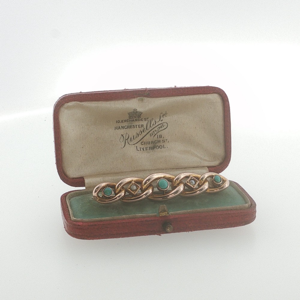 An antique 9ct rose gold bar brooch with curb links set with turquoise cabochons and seed pearls resting in a fitted presentation box.