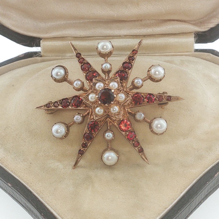 A 9ct yellow gold starburst brooch set with a central round garnet, surrounded by radiating point rays set with smaller garnets and seed pearls.