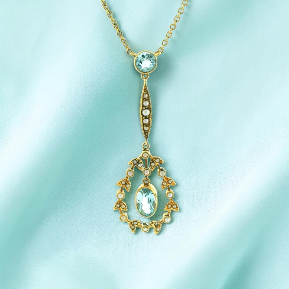 An Edwardian 15ct gold lavalier necklace featuring a round aquamarine top stone, a seed-pearl-set vertical bar, and a floral garland pendant surrounding a suspended oval aquamarine.
