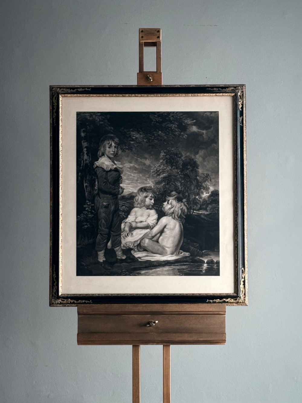 A framed monochrome antique mezzotint portrait depicting three children near a stream, housed within a dark painted frame with gilded corner accents.