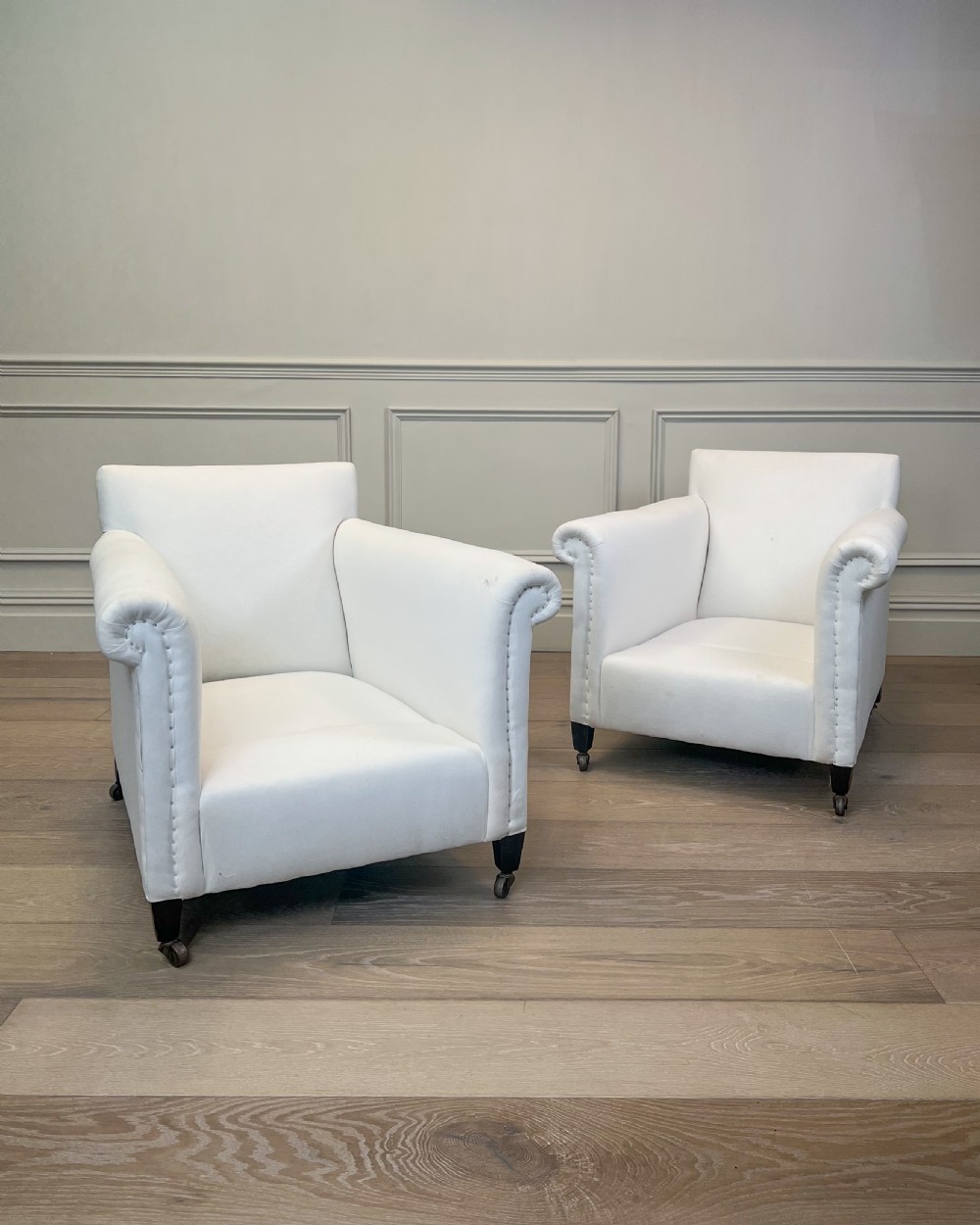 A pair of late nineteenth-century armchairs upholstered in white fabric featuring scroll arms, detail studding, and dark wooden legs with brass castors.