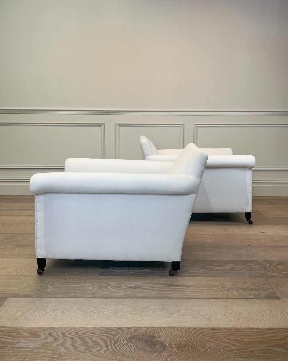 A pair of late nineteenth-century armchairs upholstered in white fabric featuring scroll arms, detail studding, and dark wooden legs with brass castors. View II