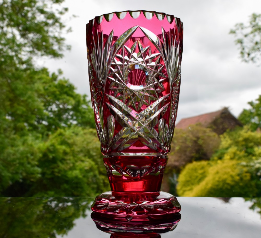 A cut-to-clear cranberry red crystal vase featuring starburst geometric cuts and a scalloped rim on a circular pedestal foot.