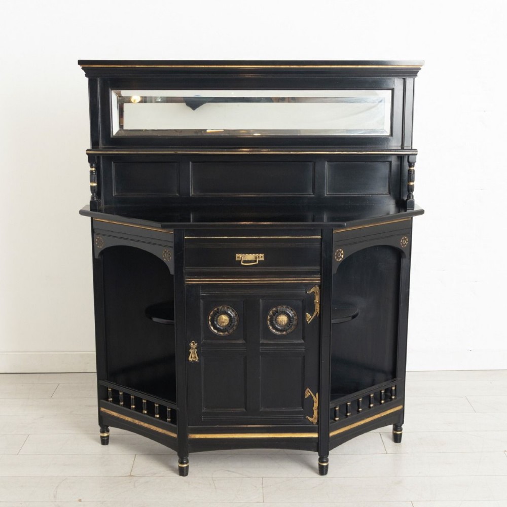 An ebonised black wood cabinet featuring gilded highlights, a mirrored top tier, open side alcoves with turned spindles, and a central cupboard door with carved circular motifs.