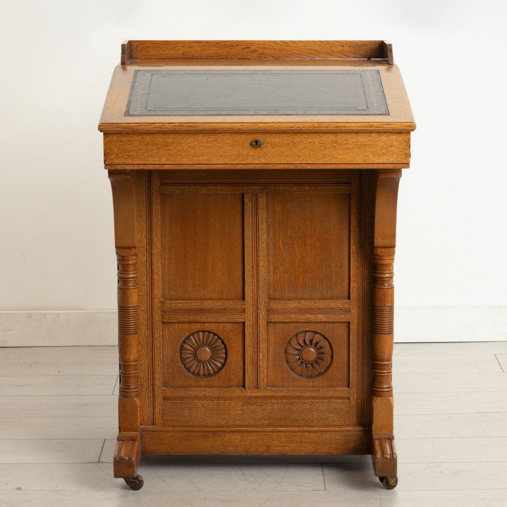 An Aesthetic Movement oak davenport desk featuring a sloped writing surface inset with green leather, carved roundels on the front panel, turned columns, and brass castors.