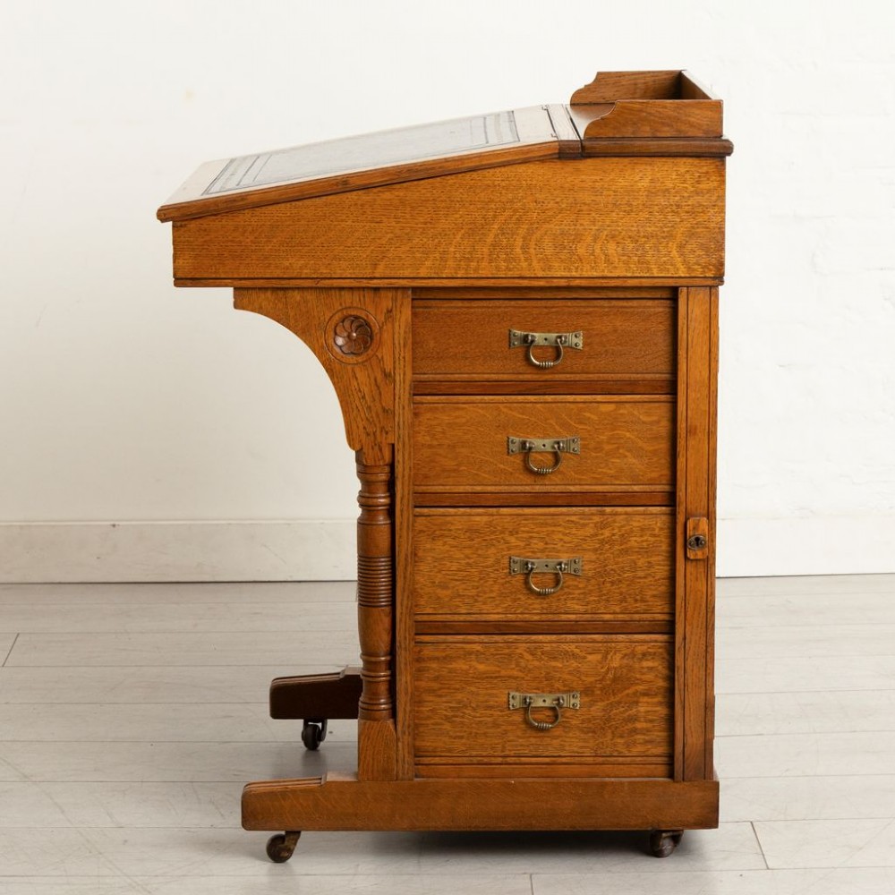 An Aesthetic Movement oak davenport desk featuring a sloped writing surface inset with green leather, carved roundels on the front panel, turned columns, and brass castors. View II