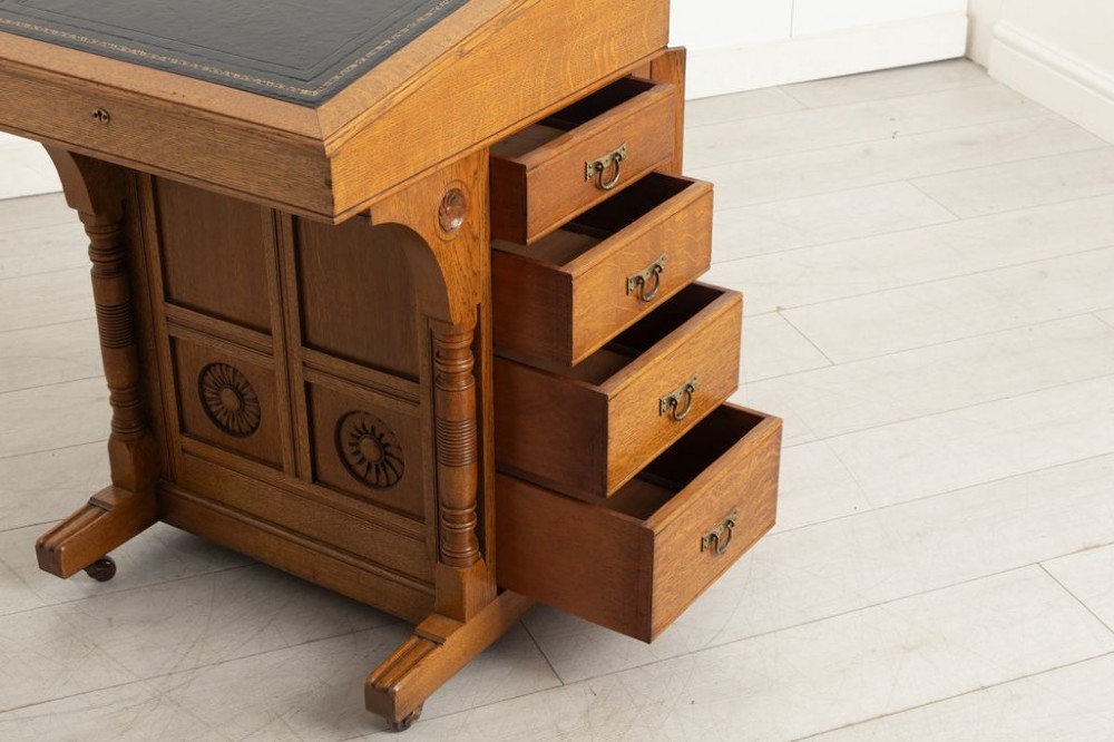 An Aesthetic Movement oak davenport desk featuring a sloped writing surface inset with green leather, carved roundels on the front panel, turned columns, and brass castors. View III