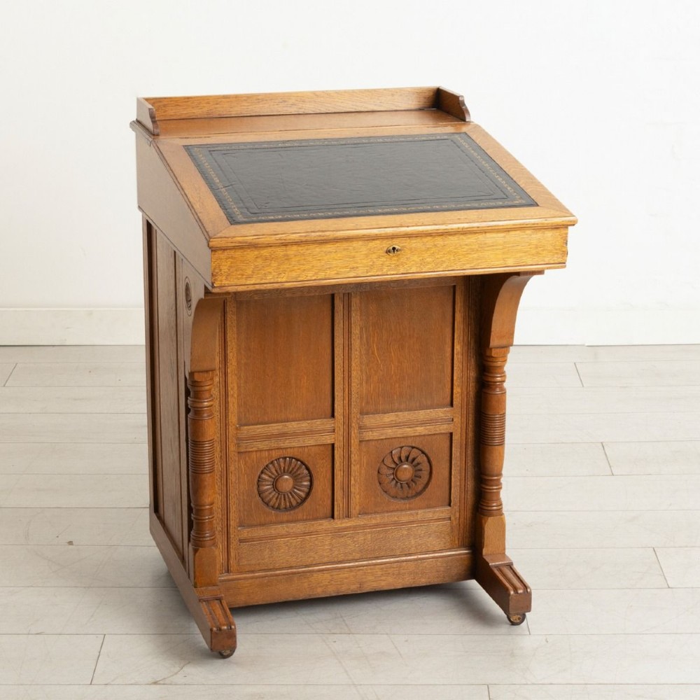 An Aesthetic Movement oak davenport desk featuring a sloped writing surface inset with green leather, carved roundels on the front panel, turned columns, and brass castors. View IV