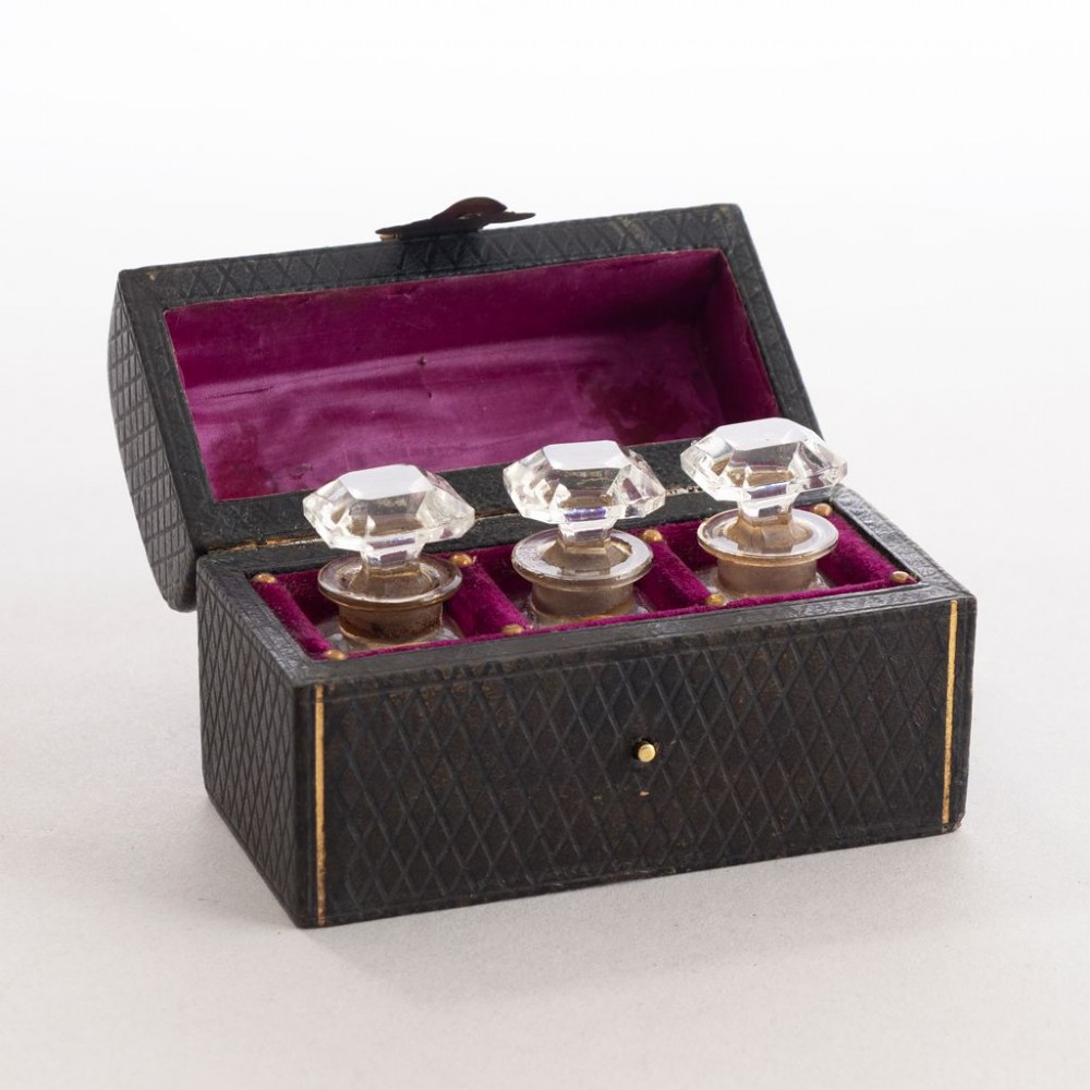 An open black leather box lined with purple velvet containing three clear glass perfume bottles with octagonal faceted stoppers.