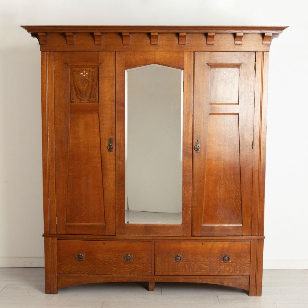 A large oak three-door Arts and Crafts wardrobe featuring a central arched mirror flanked by stylized inlaid panels over two lower drawers.