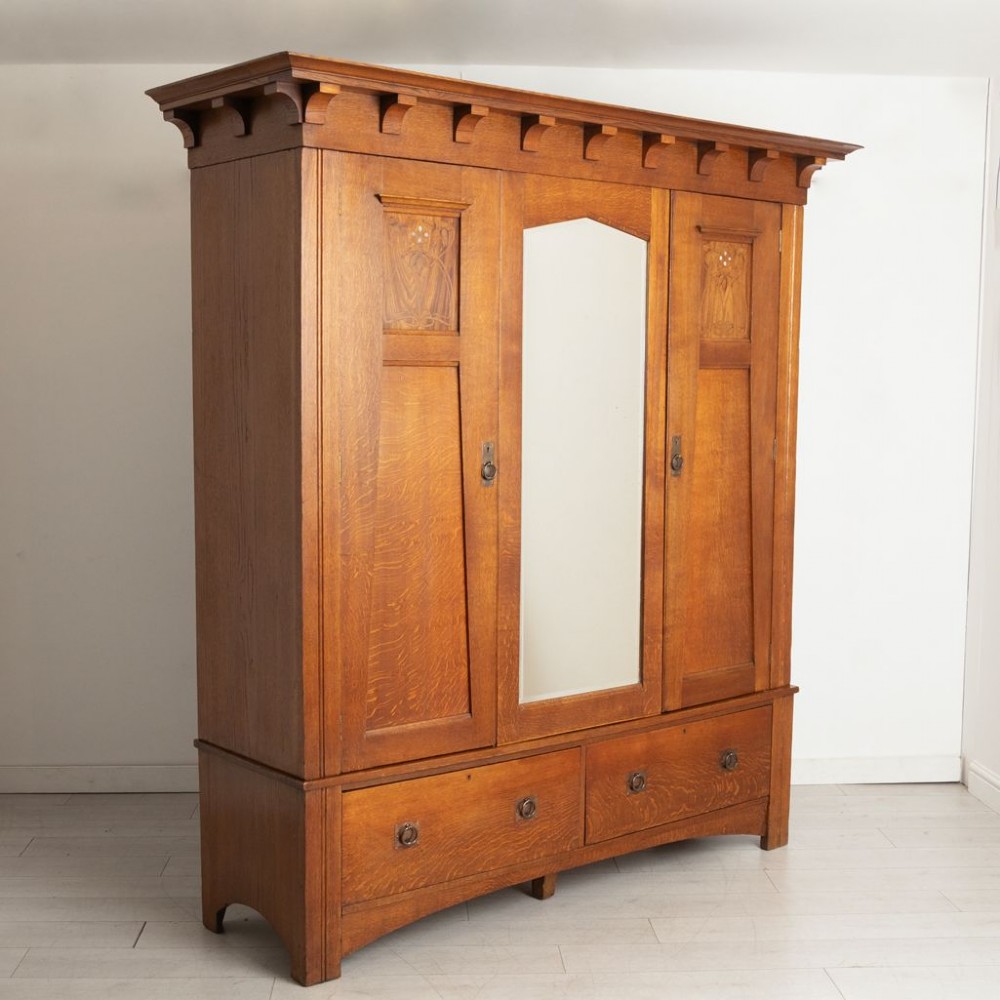 A large oak three-door Arts and Crafts wardrobe featuring a central arched mirror flanked by stylized inlaid panels over two lower drawers. View II