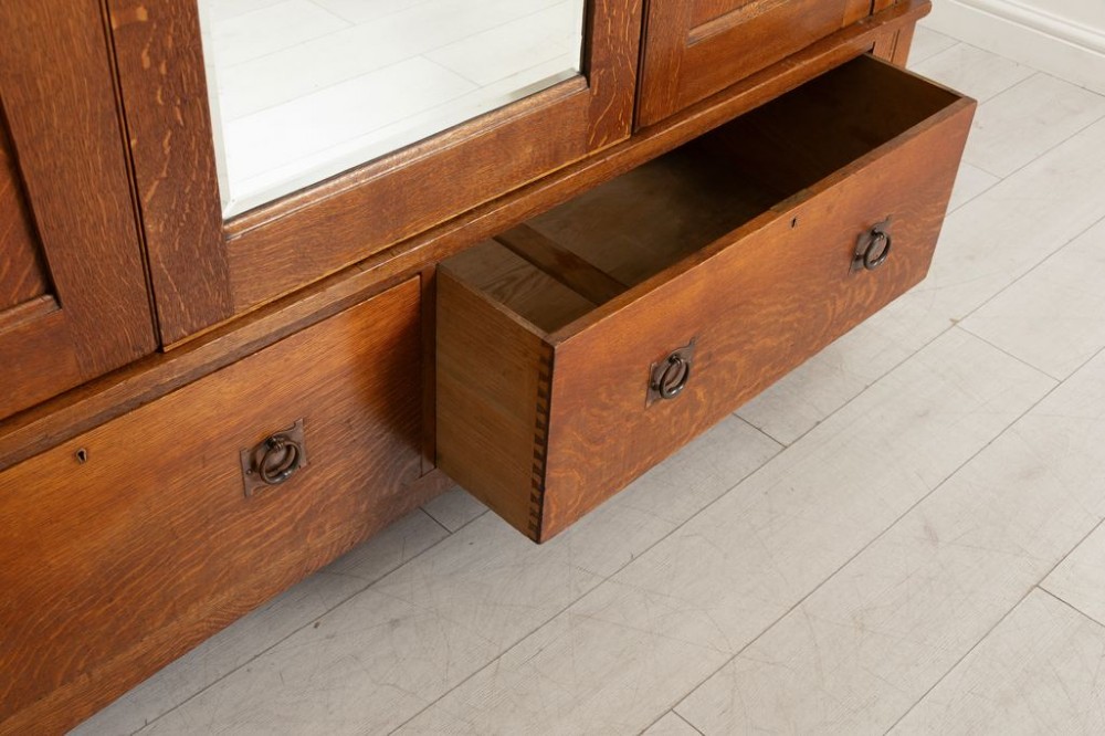A large oak three-door Arts and Crafts wardrobe featuring a central arched mirror flanked by stylized inlaid panels over two lower drawers. View III