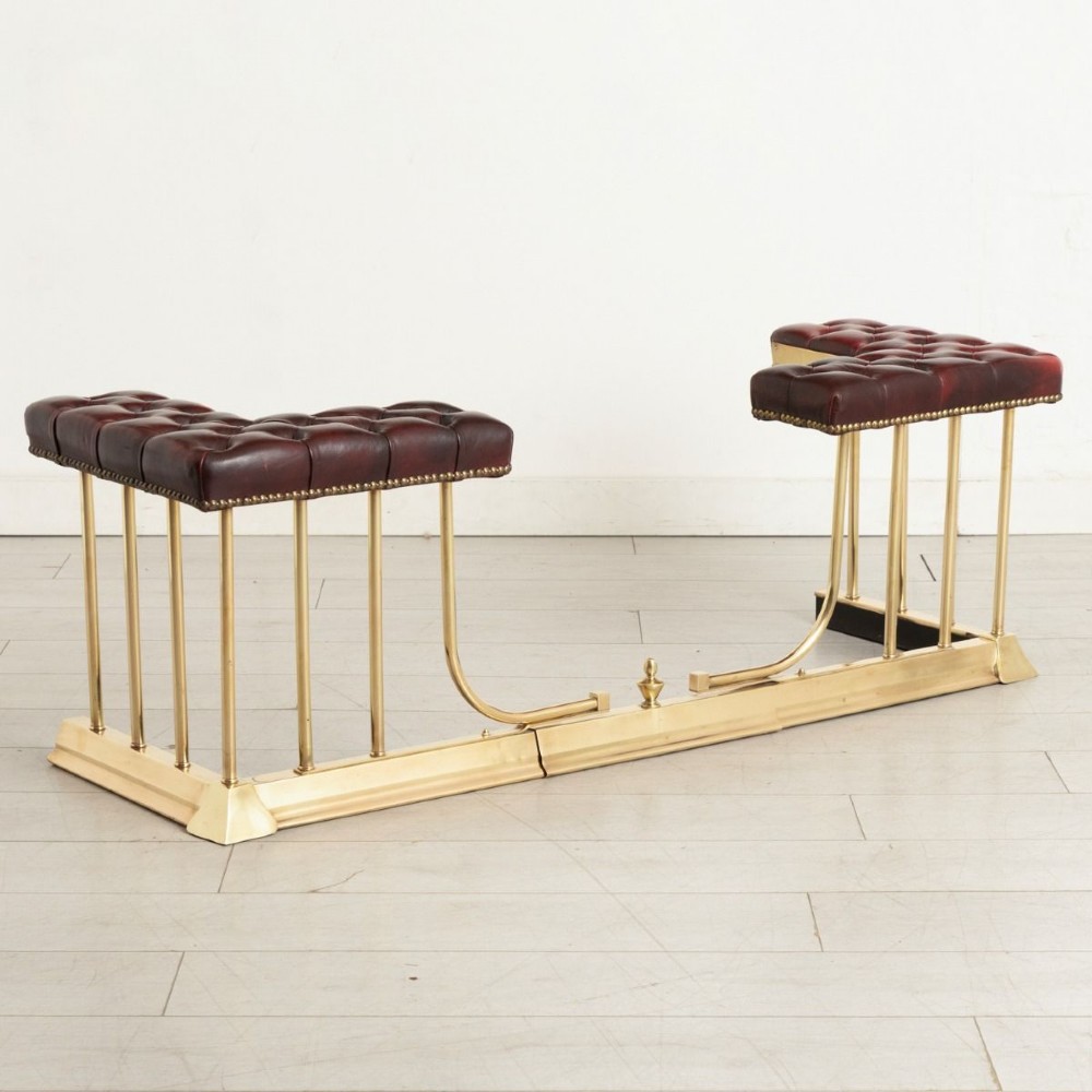 An Edwardian polished brass club fender featuring twin burgundy leather buttoned seats supported by vertical brass rails on a shaped base.