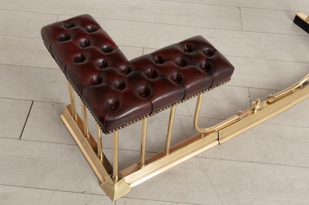 An Edwardian polished brass club fender featuring twin burgundy leather buttoned seats supported by vertical brass rails on a shaped base. View II