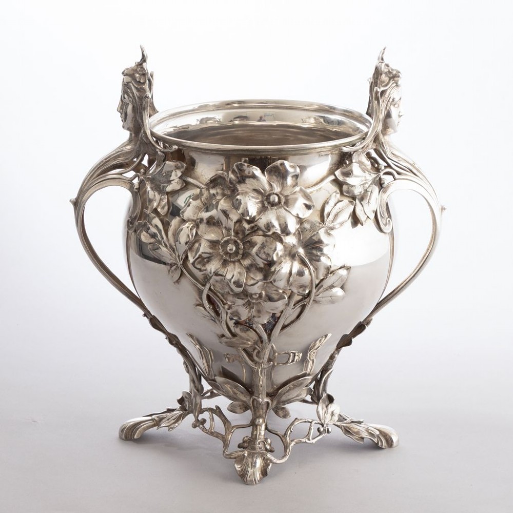 A silver-plated Art Nouveau champagne bucket featuring floral relief details, whiplash tendrils, sculpted handles with female heads, and a pierced footed base.