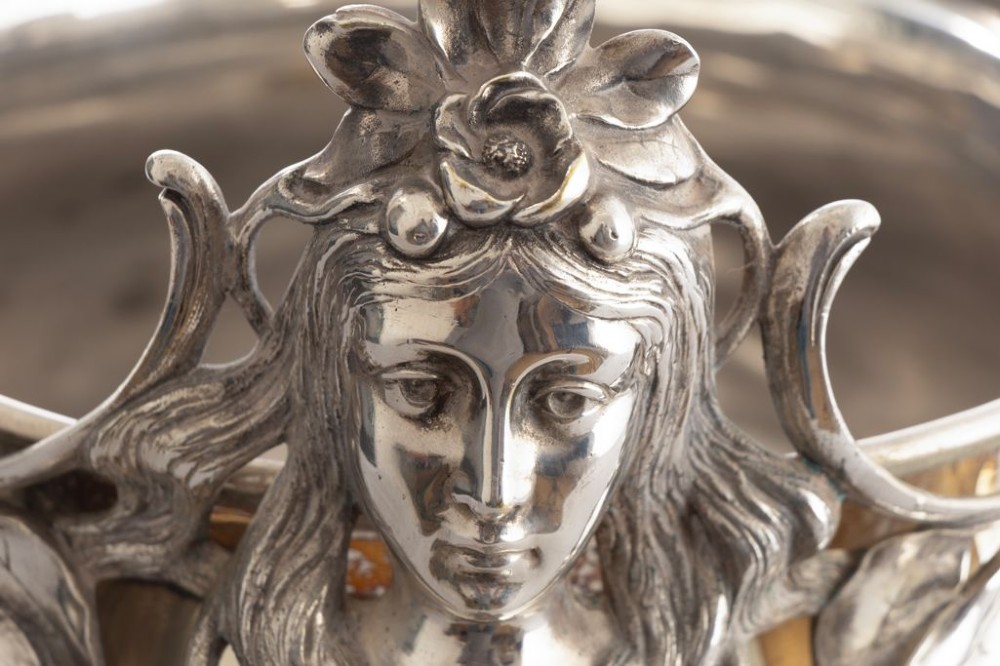 A silver-plated Art Nouveau champagne bucket featuring floral relief details, whiplash tendrils, sculpted handles with female heads, and a pierced footed base. View V