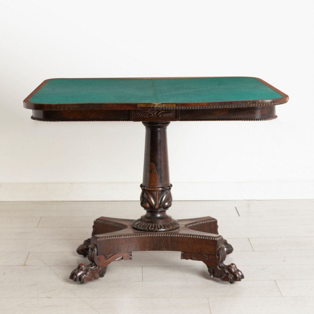 An antique mahogany gaming table featuring an unfolded green baize playing surface supported by a central carved pedestal stem and quadform base.