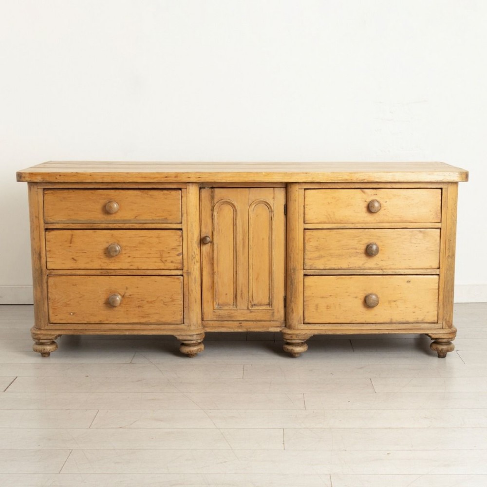A 19th-century honey pine dresser base featuring six side drawers flanking a central arched cupboard door resting on turned bun feet.