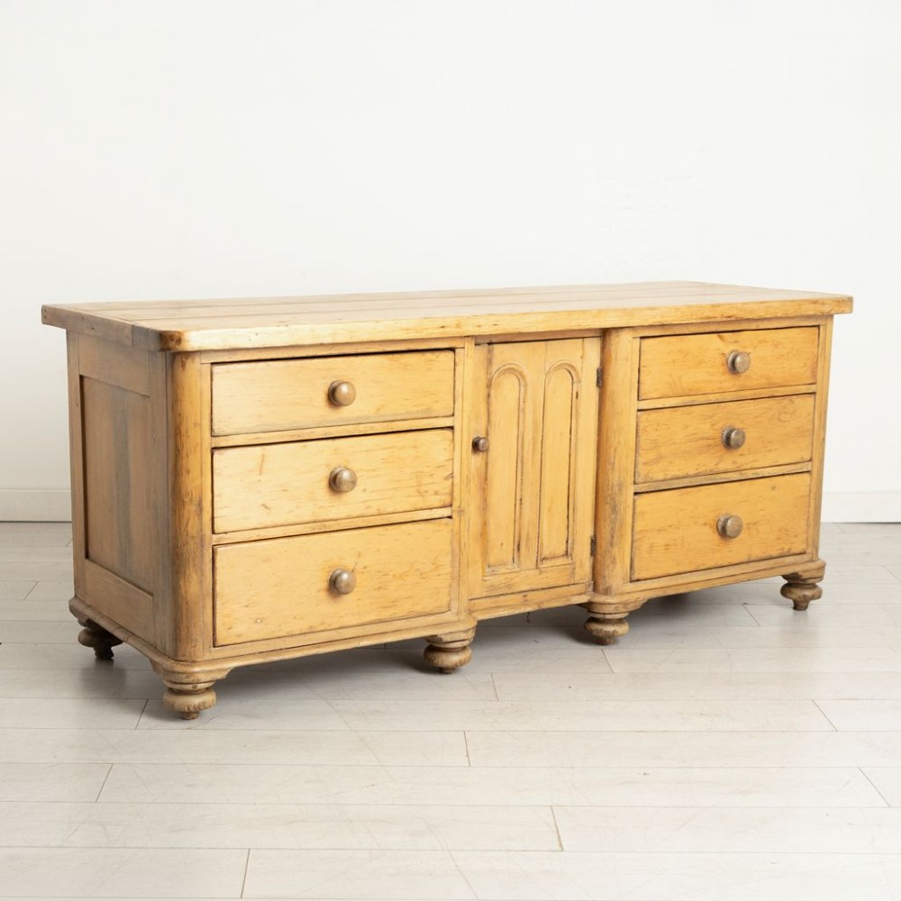 A 19th-century honey pine dresser base featuring six side drawers flanking a central arched cupboard door resting on turned bun feet. View II
