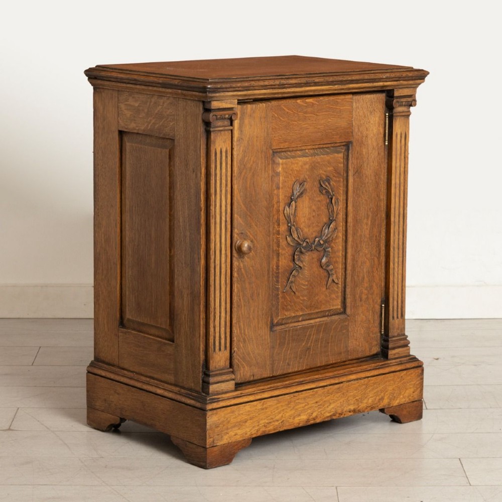 A late nineteenth-century oak side cupboard featuring a single panelled door decorated with a carved laurel wreath motif and flanked by fluted pilasters.