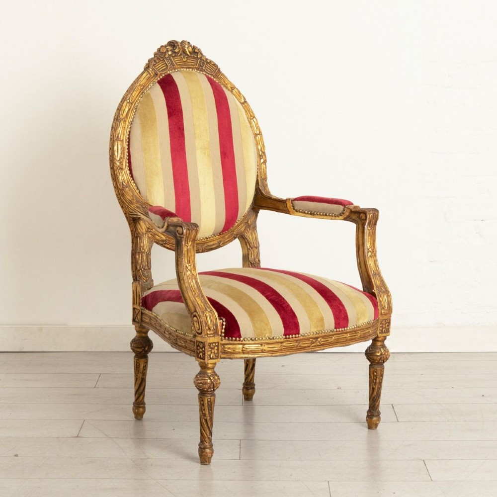 A French Louis XVI style open armchair featuring a carved gold giltwood frame, oval backrest, fluted legs, and red and cream striped velvet upholstery. View II