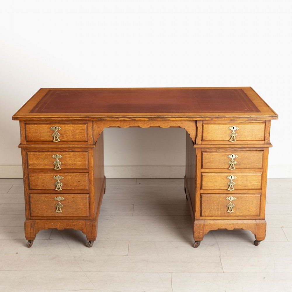 An Arts and Crafts golden oak pedestal desk featuring an inset leather writing surface, twin pedestals with brass handles, and castors.