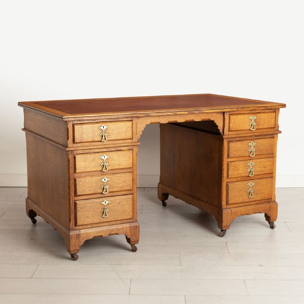 An Arts and Crafts golden oak pedestal desk featuring an inset leather writing surface, twin pedestals with brass handles, and castors. View II
