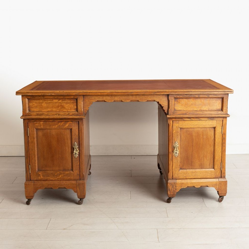 An Arts and Crafts golden oak pedestal desk featuring an inset leather writing surface, twin pedestals with brass handles, and castors. View III