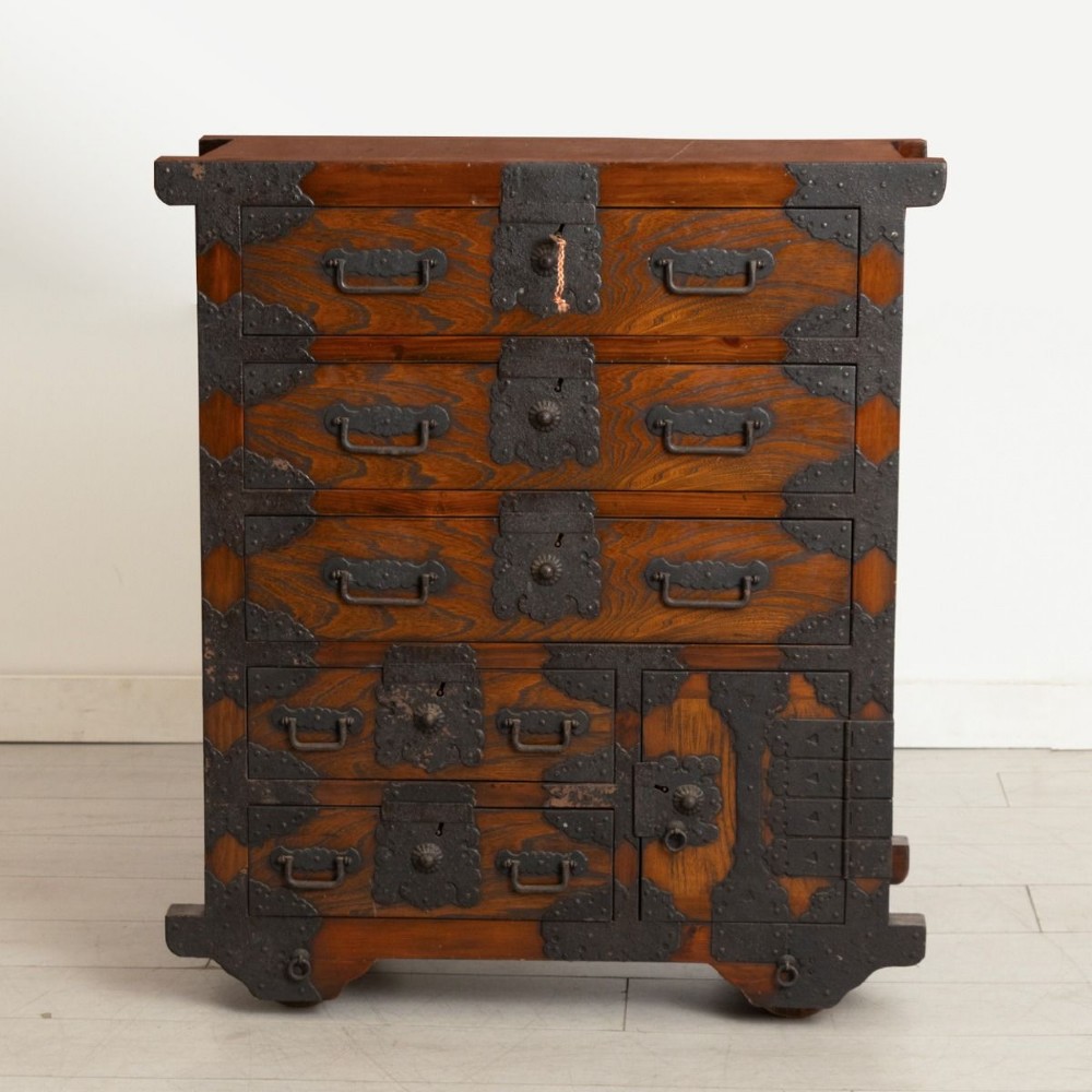 An antique Japanese zelkova wood chest of drawers featuring dark iron corner brackets, lock plates, drop handles, and integrated wooden wheels at the base.