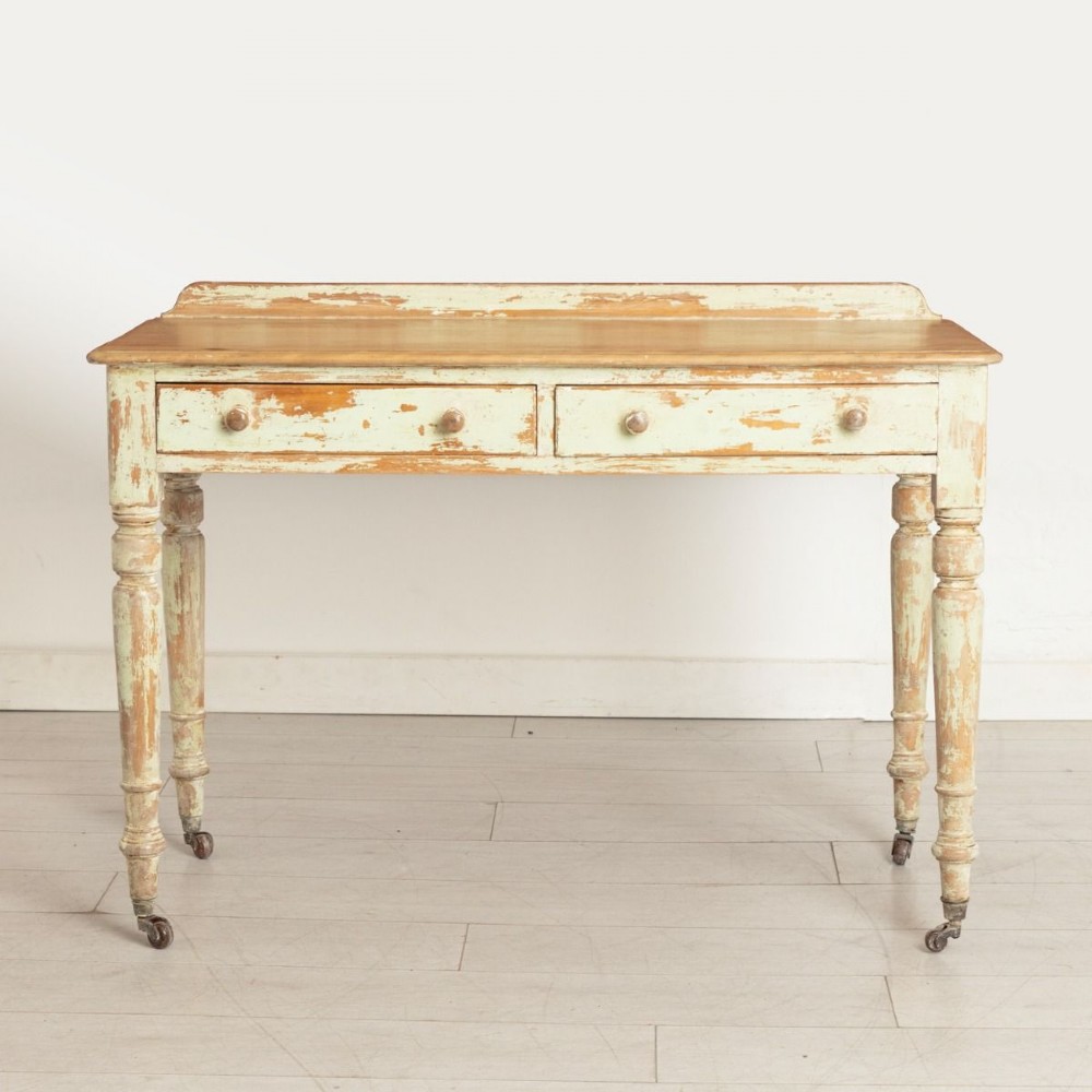 A 19th-century birch writing table with a polished natural timber top, two drawers with wooden knobs, distressed off-white painted turned legs, and brass castors.