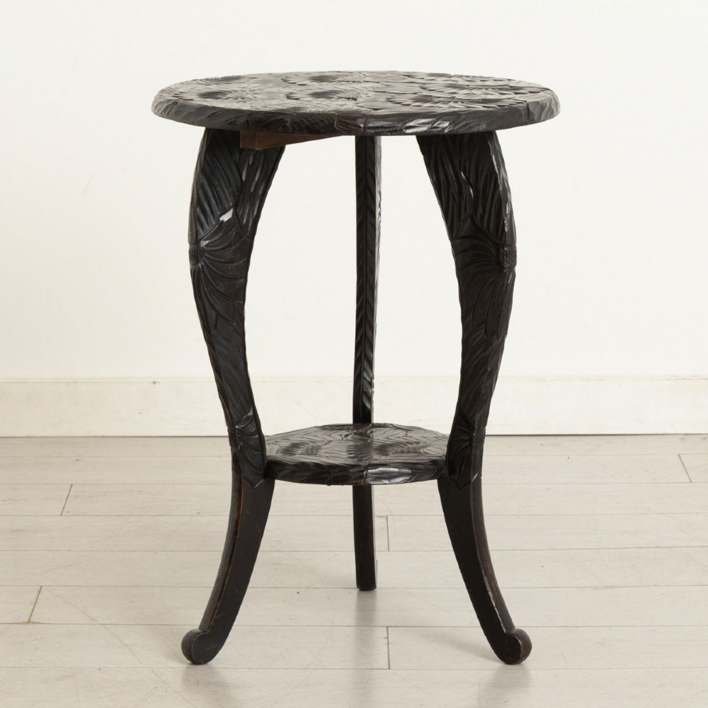 A dark-stained wooden side table featuring a circular top and three outward-curving legs detailed with relief floral carvings and a small lower shelf.