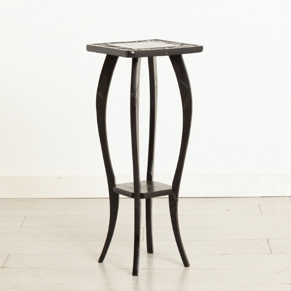 A tall ebonised wooden plant stand featuring carved floral detailing on the square top surface, slender curved legs, and an lower tier shelf.