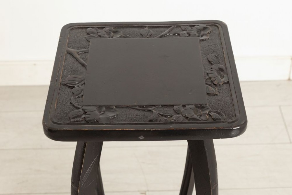 A tall ebonised wooden plant stand featuring carved floral detailing on the square top surface, slender curved legs, and an lower tier shelf. View II