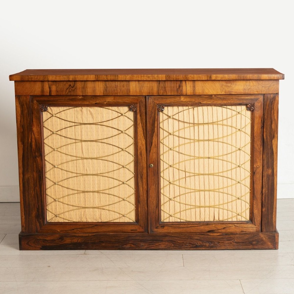 An antique mid-nineteenth century rosewood side cabinet featuring two doors with brass wire grilles over pleated fabric panels and a plinth base.
