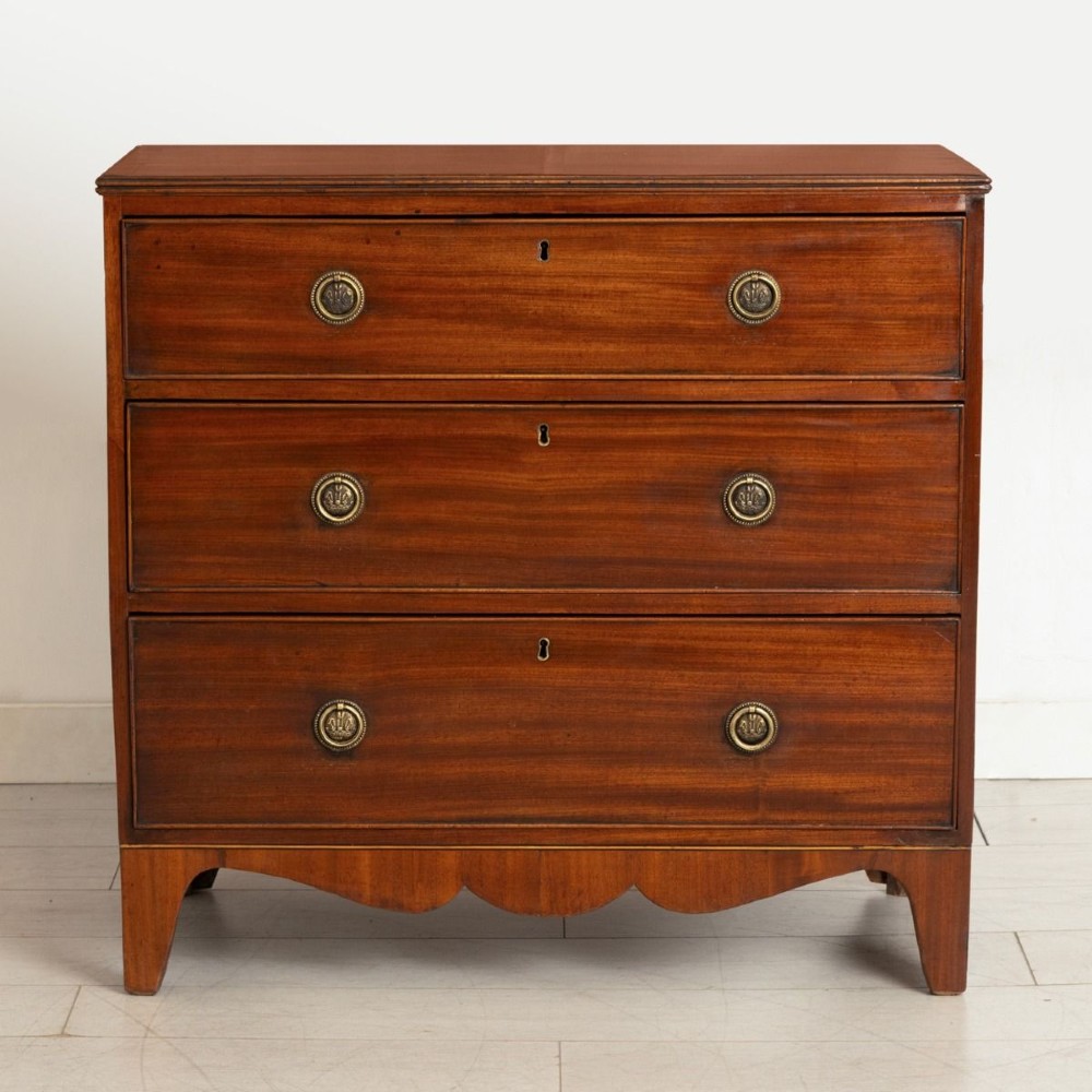A Georgian mahogany chest of three long drawers featuring brass ring handles, escutcheons, and a scalloped apron resting on bracket feet.
