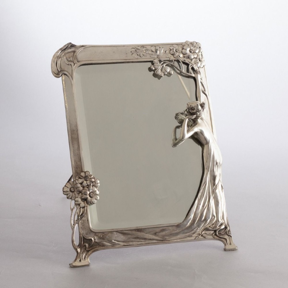 An Art Nouveau pewter easel-backed mirror featuring a sculptured maiden figure and relief floral motifs around a rectangular glass frame.