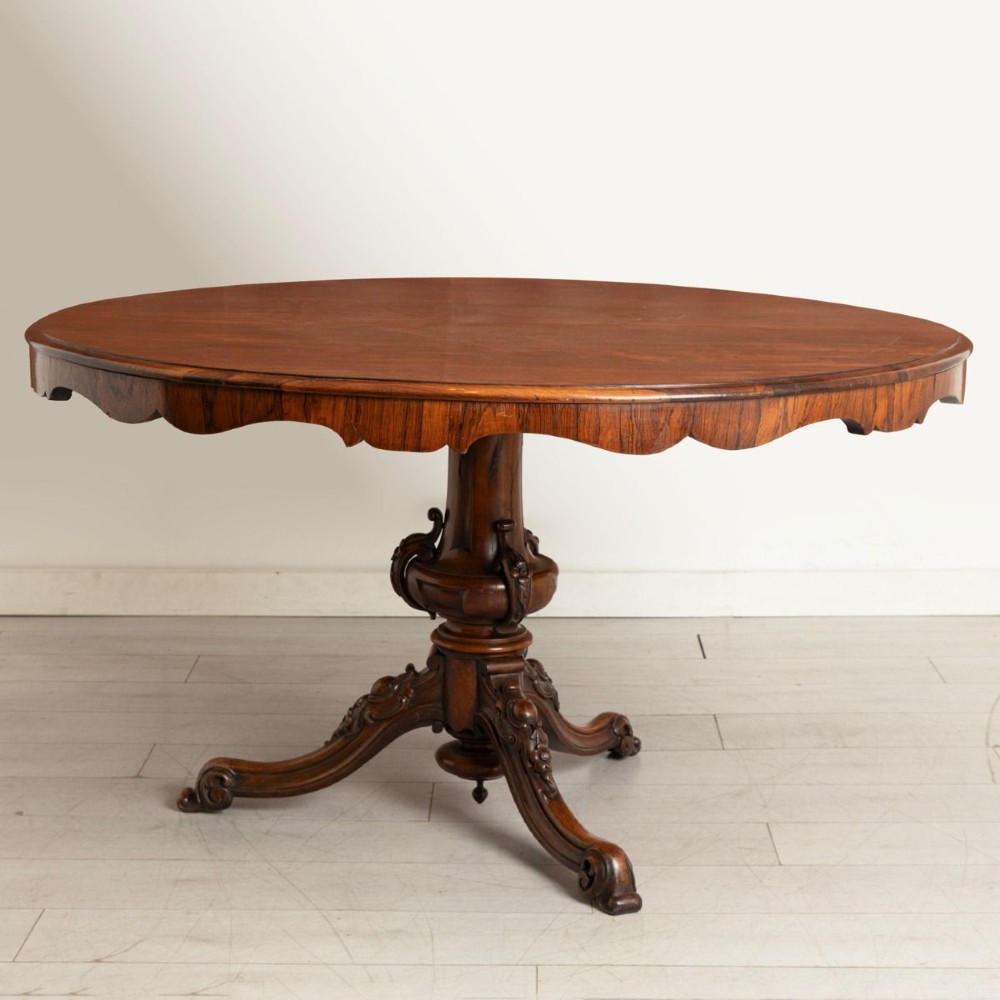 A Victorian rosewood tilt-top dining table featuring an oval top with a scalloped apron, supported on a carved pedestal column with scroll tripod legs.