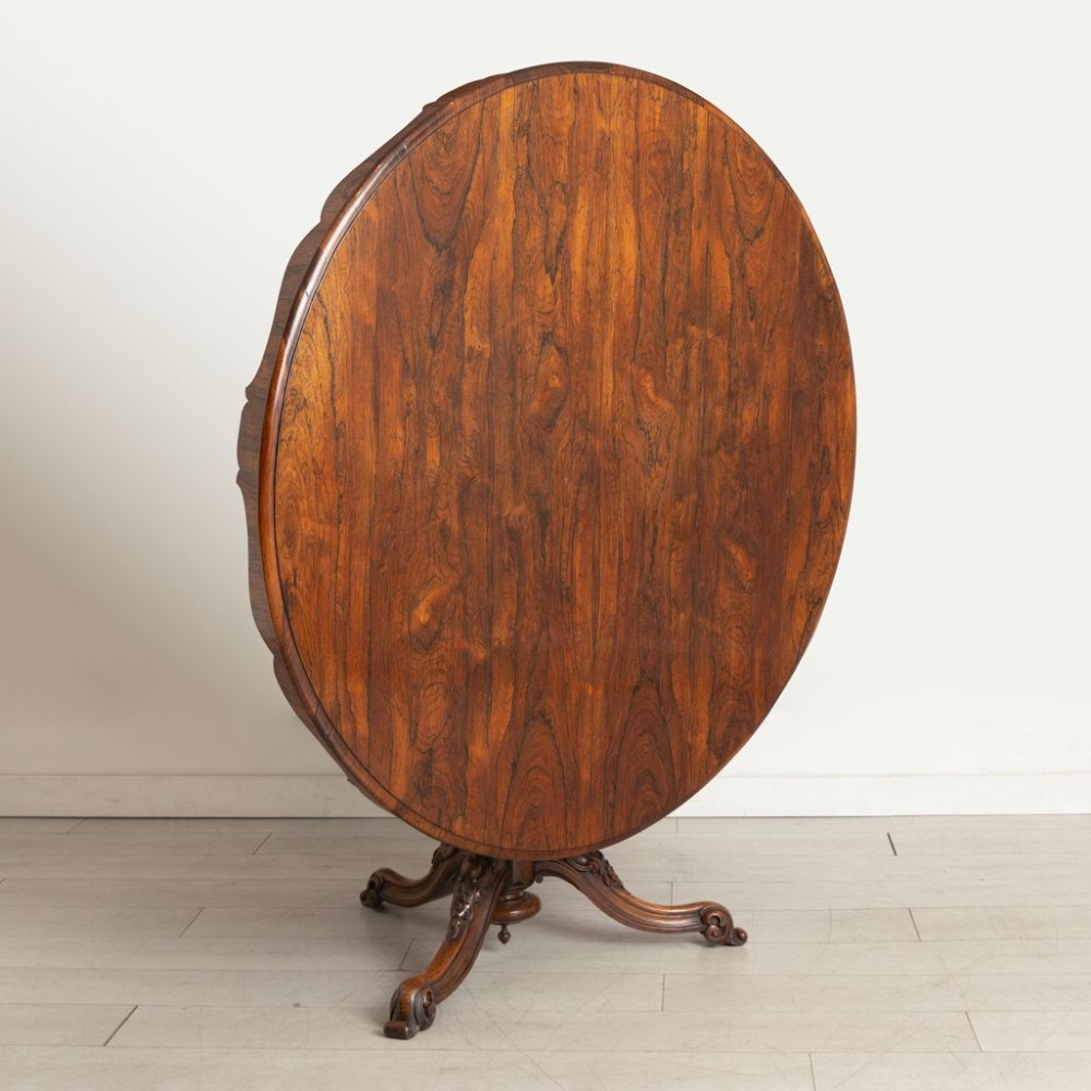 A Victorian rosewood tilt-top dining table featuring an oval top with a scalloped apron, supported on a carved pedestal column with scroll tripod legs. View II