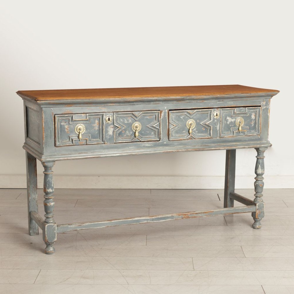 An antique oak dresser base featuring a natural wooden top, distressed grey painted finish, geometric moulded drawers with brass handles, and turned legs with a lower stretcher. View II