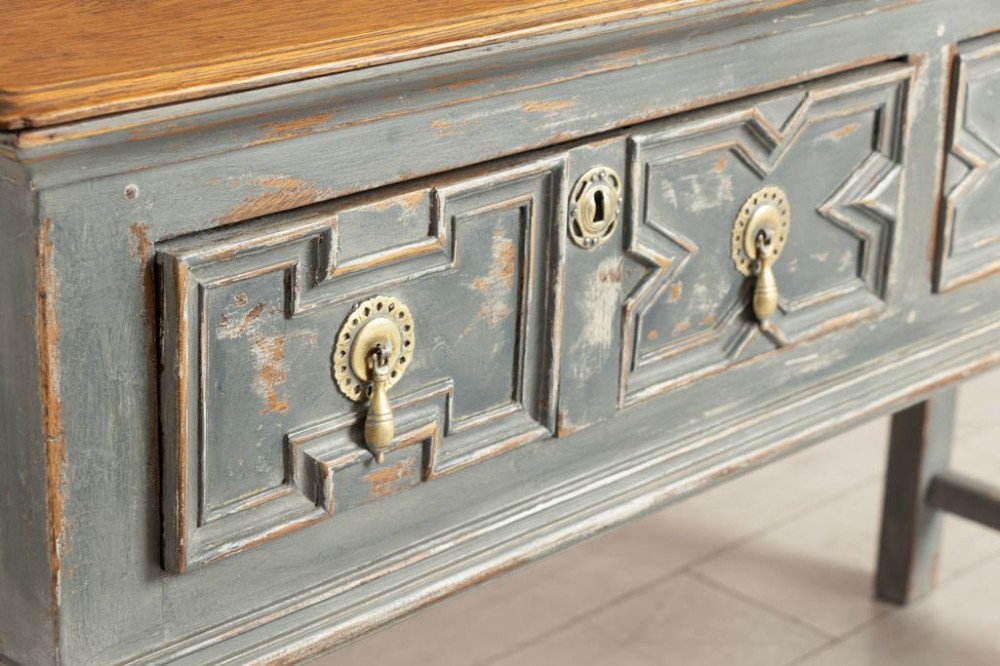 An antique oak dresser base featuring a natural wooden top, distressed grey painted finish, geometric moulded drawers with brass handles, and turned legs with a lower stretcher. View III