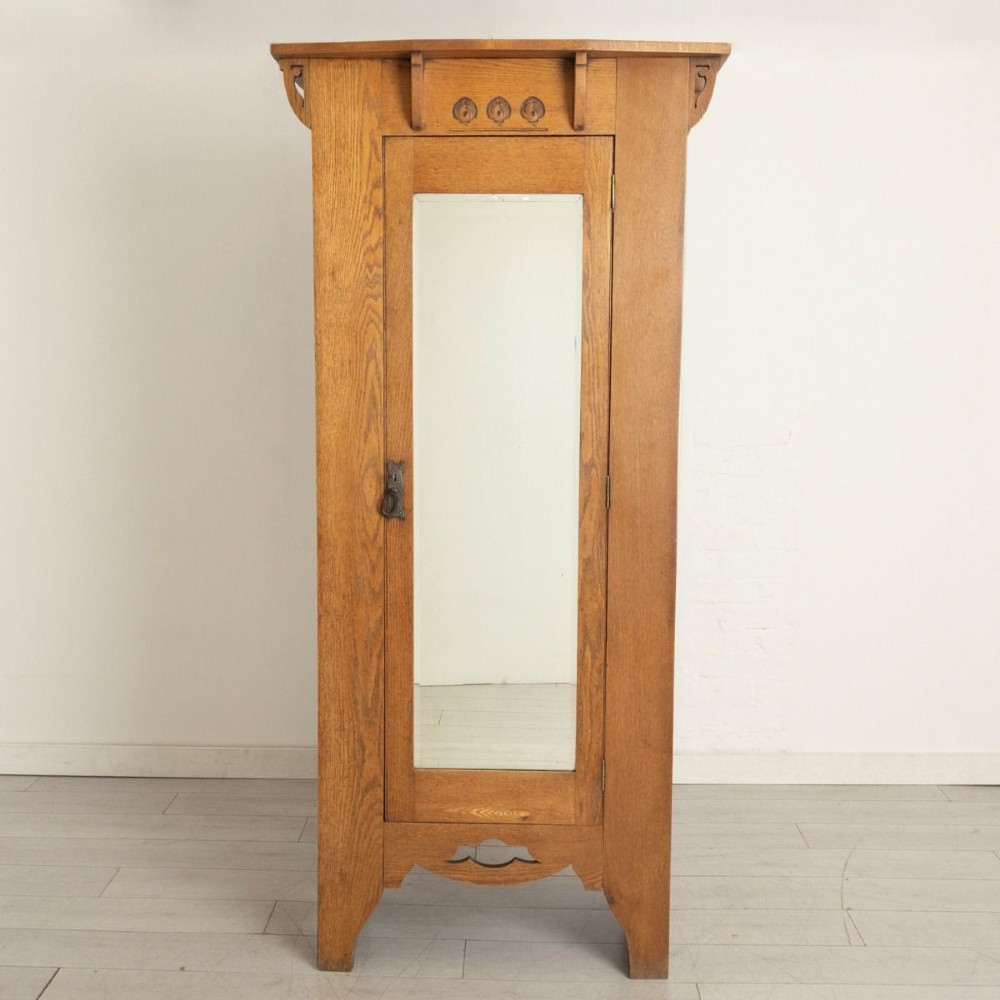 An Arts and Crafts solid oak single door wardrobe featuring a central full-length mirror, carved decorative details beneath an overhanging cornice, and a shaped apron.